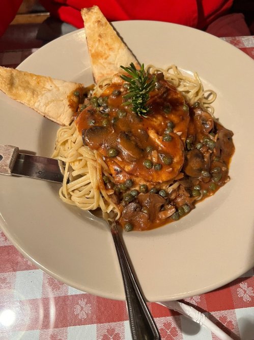 A plate of pasta topped with chicken, mushroom sauce, capers, and garnished with a sprig of rosemary, served with bread.