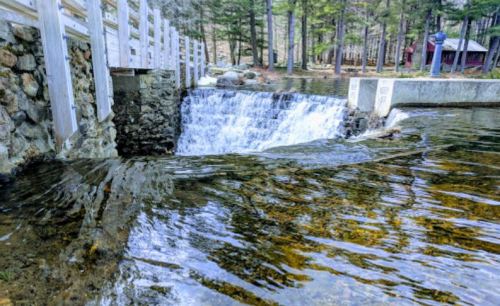 A serene waterfall cascades over a stone barrier, surrounded by trees and a calm body of water.