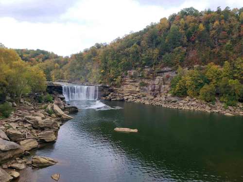 A serene waterfall cascades into a calm river, surrounded by colorful autumn foliage and rocky terrain.