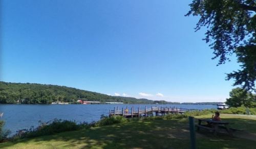 A serene lakeside view with a wooden dock, green hills, and a clear blue sky. A person sits at a picnic table nearby.