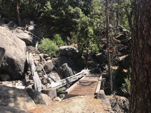 A wooden bridge crosses a rocky path surrounded by trees and boulders in a natural setting.
