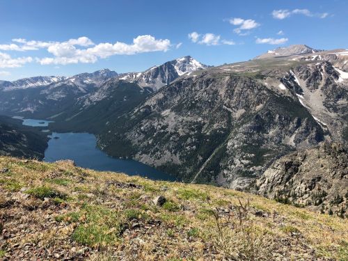 A panoramic view of mountains and a lake under a clear blue sky, showcasing rugged terrain and lush greenery.