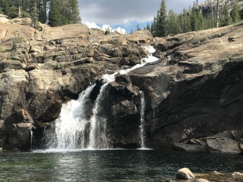 A serene waterfall cascading over rocky terrain into a clear pool, surrounded by trees and a cloudy sky.