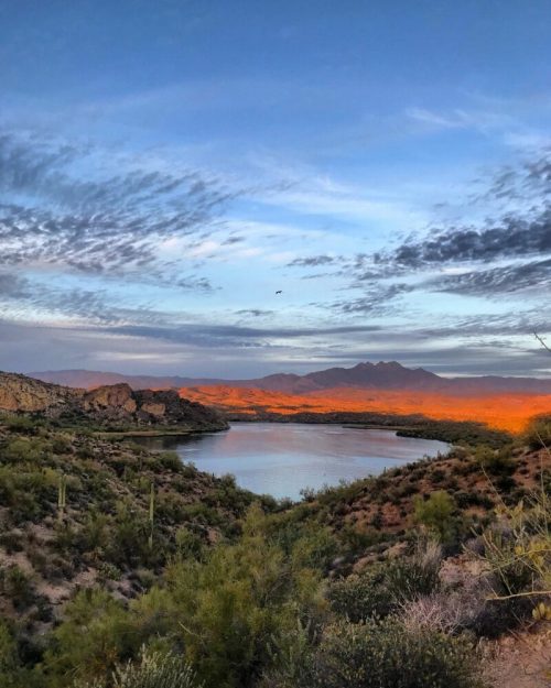 A serene landscape featuring a lake surrounded by mountains and desert vegetation under a colorful sky at sunset.