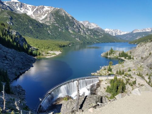 A scenic view of a dam spilling water into a serene lake, surrounded by mountains and lush greenery under a clear blue sky.