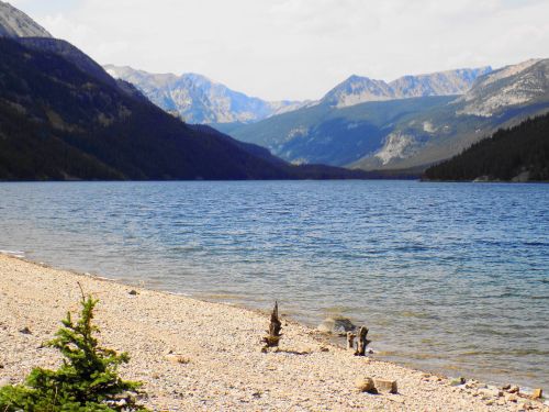 A serene lake surrounded by mountains, with a pebbly shore and a few trees in the foreground.