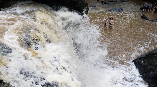 Two people stand in shallow water near a waterfall, with others enjoying the rocky shoreline in the background.