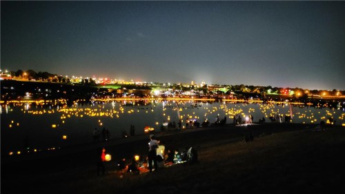 A serene night scene with glowing lanterns floating on a lake, surrounded by city lights and people enjoying the view.