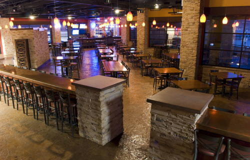 A spacious restaurant interior with stone walls, wooden tables, and pendant lighting, featuring a bar area in the background.