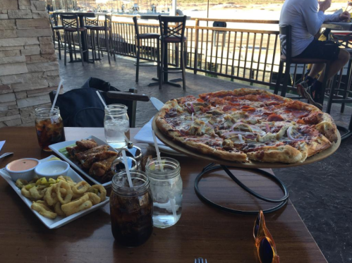 A large pizza on a wooden table with wings, onion rings, and drinks, set in a restaurant with a view.
