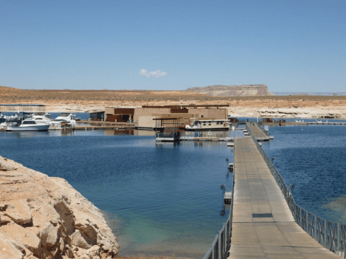 A marina with boats docked, featuring a long pier extending into calm blue water under a clear sky.