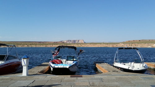 Two boats docked at a marina on a calm lake, with a clear blue sky and distant hills in the background.