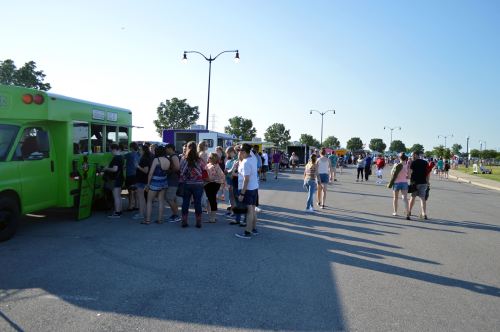 A crowd gathers around food trucks at an outdoor event on a sunny day. Trees and streetlights line the background.