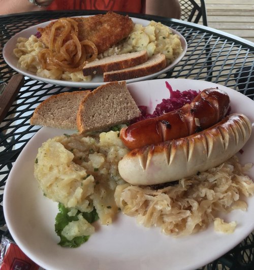 A plate of sausages, mashed potatoes, sauerkraut, red cabbage, bread, and fried onions on a table.