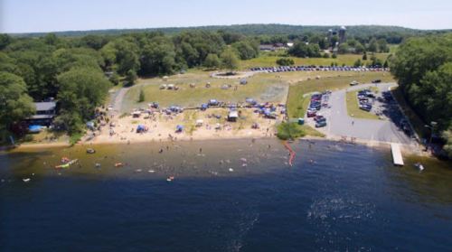 Aerial view of a beach with people, tents, and cars parked nearby, surrounded by trees and water.