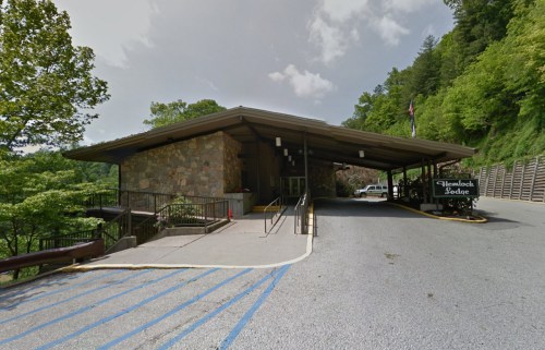 Exterior view of a stone lodge building surrounded by greenery, with a sloped driveway and parking area.