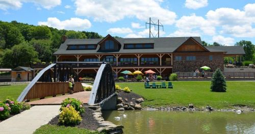 A scenic view of a lodge with a bridge over a pond, surrounded by greenery and colorful umbrellas on a sunny day.