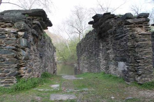 Ruins of stone walls with a pathway leading to a calm body of water, surrounded by trees and greenery.