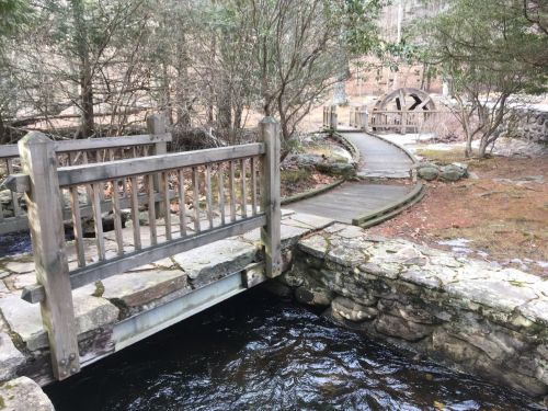 A wooden bridge over a stream, with a winding path and trees in the background.