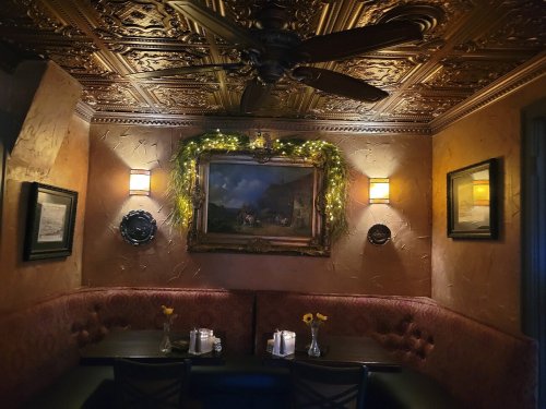 Cozy restaurant corner with ornate ceiling, warm lighting, and a painting, featuring a booth with flowers on the table.