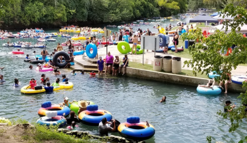 A busy river scene with people floating on colorful tubes, swimming, and relaxing along the shore. Lush greenery surrounds the area.