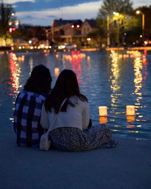 Two people sit by a calm water body at dusk, surrounded by glowing lanterns and city lights reflecting on the surface.