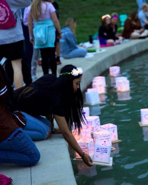 A person leans over a pond, placing a glowing lantern on the water among others, with a group of people in the background.