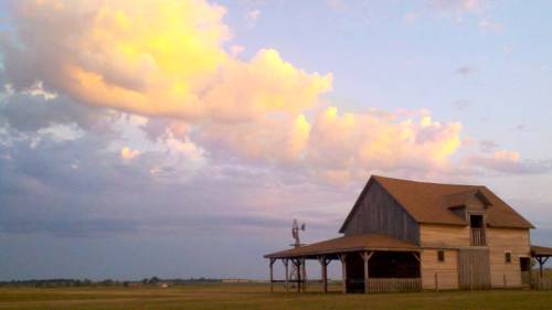 A rustic barn stands in a field under a colorful sky with fluffy clouds at sunset.