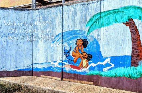 Colorful mural depicting two girls surfing with a dog, welcoming visitors to Historic Old Town Hanapepe, home of Lilo & Stitch.