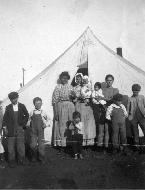 A historical black-and-white photo of a group of adults and children standing in front of a large tent.