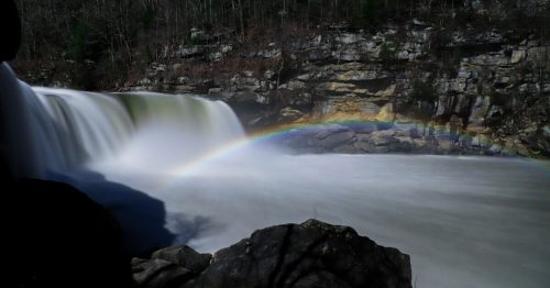 A waterfall cascades over rocks, creating a misty rainbow under a starry night sky.