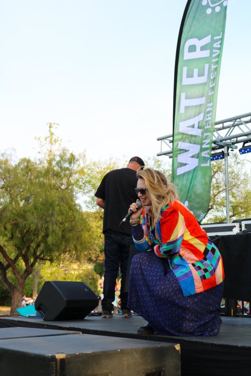 A performer in a colorful jacket sings on stage at the Water Lantern Festival, with trees and a crowd in the background.