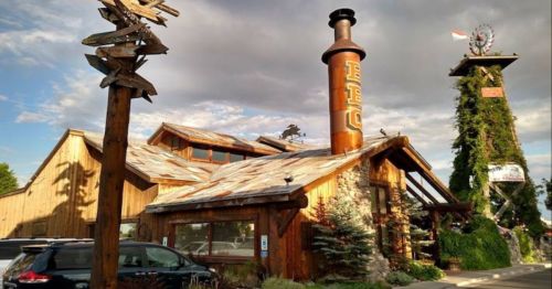 A rustic wooden building with a tall chimney, surrounded by trees and decorative signs, under a cloudy sky.