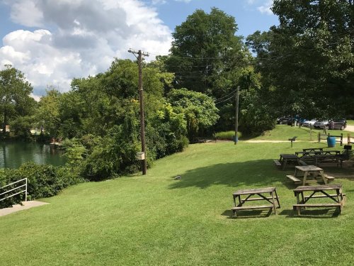 A grassy area with picnic tables near a river, surrounded by trees and under a partly cloudy sky.