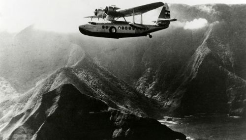 A vintage seaplane flying over rugged mountains and a coastline, with clouds in the background.
