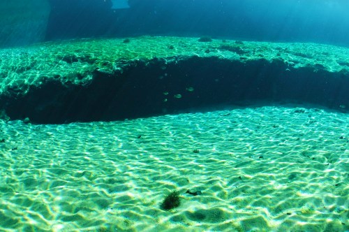 Underwater scene with sandy bottom, sunlight filtering through water, and small fish swimming in a clear blue environment.