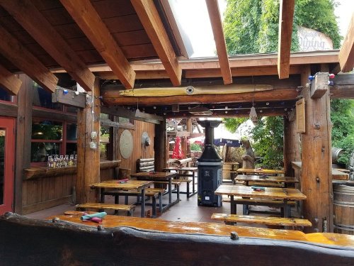 Cozy outdoor dining area with wooden tables, a black stove, and greenery overhead. Warm, rustic atmosphere.