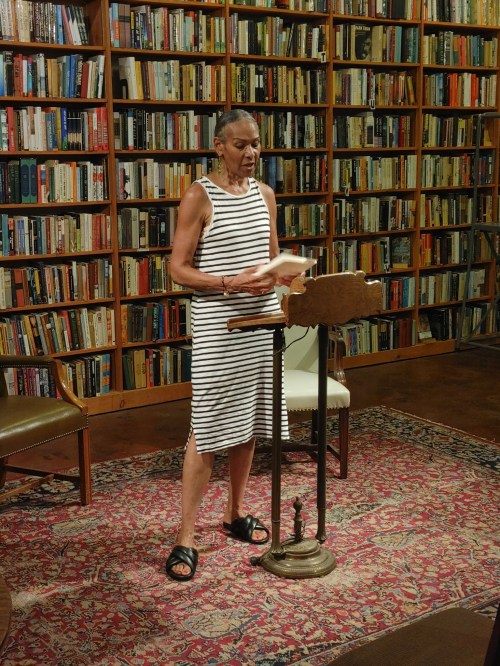 A woman in a striped dress stands at a lectern, reading in a library filled with books. A rug and chair are nearby.