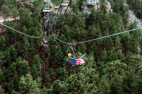 A person in a harness glides on a zip line over a dense forest, with rocky cliffs in the background.
