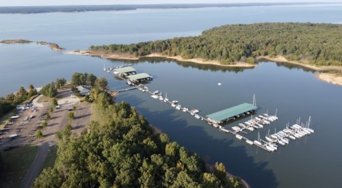 Aerial view of a marina with boats docked, surrounded by trees and calm water. Parking area visible nearby.