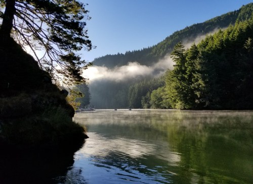 A serene lake surrounded by misty mountains and lush green trees under a clear blue sky.