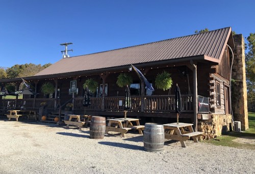 A rustic wooden building with a metal roof, featuring outdoor seating and hanging plants, set against a clear blue sky.