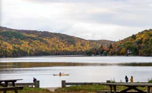 A serene lake surrounded by autumn-colored hills, with a person kayaking and others walking along the shore.