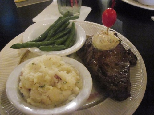 A plate featuring a grilled steak, mashed potatoes, green beans, and a cherry tomato garnish on top of a butter pat.