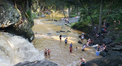 A scenic river with a waterfall, surrounded by people enjoying the water and nature in a lush, green setting.
