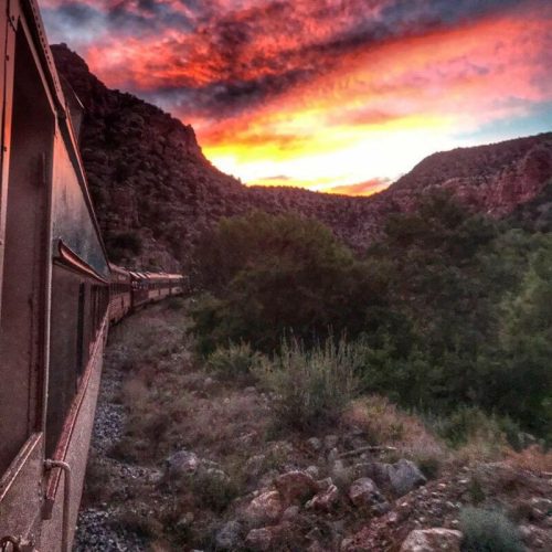 A train travels along a rocky path with vibrant sunset colors illuminating the sky and surrounding mountains.