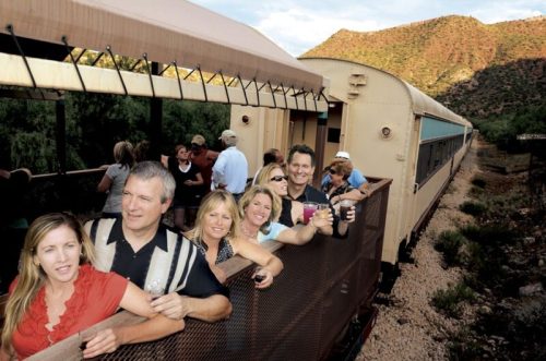 A group of people enjoying a scenic train ride, smiling and holding drinks while leaning out of the train car.