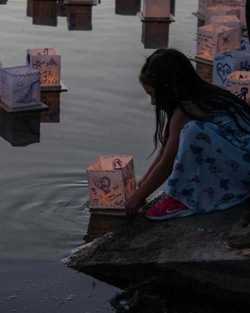 A girl in a dress places a decorated lantern into a calm body of water at dusk.