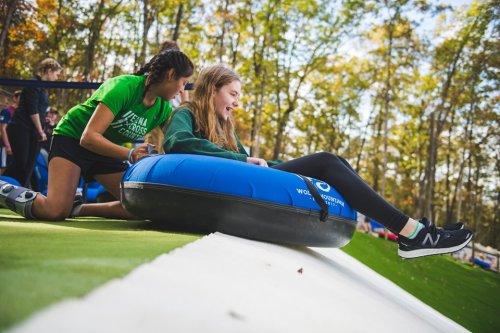 A girl sits on a blue tube, ready to slide down, while another girl assists her in a fun outdoor setting.