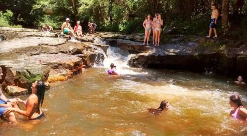 A group of people enjoying a sunny day by a natural swimming area with rocks and flowing water.